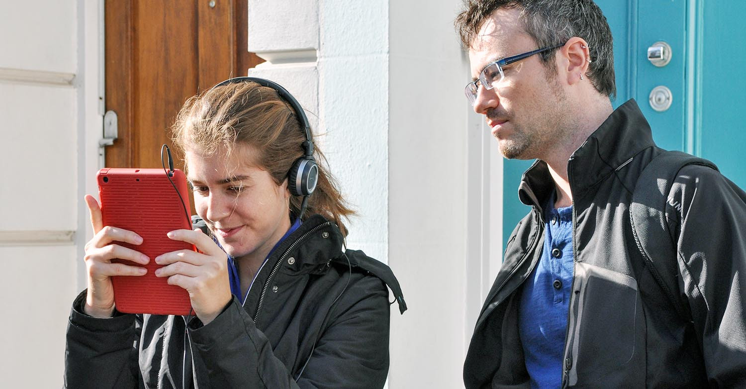 Photo of a female student testing software using an iPad and headphones while being observed by a male student