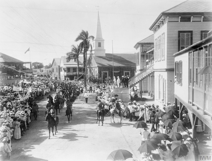 Black and white photograph taken in Jamaica and showing officers on horseback leading a parade of troops past a cheering crowd